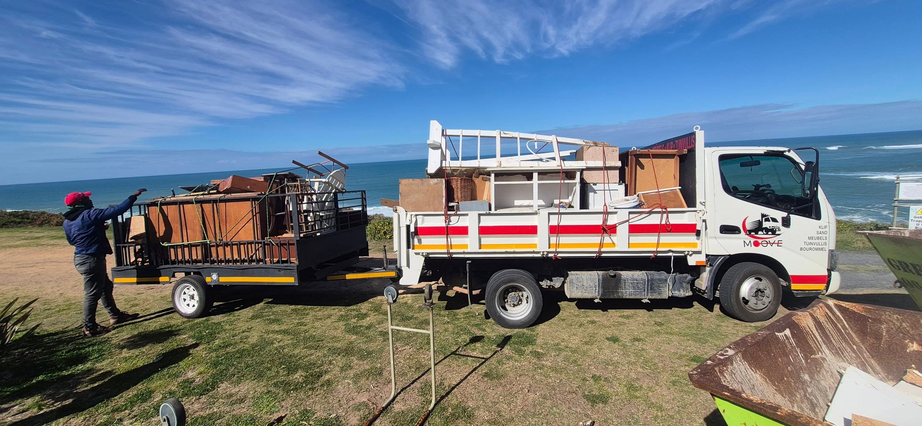 White truck and trailer loaded with furniture parked on a grassy cliff overlooking the ocean.